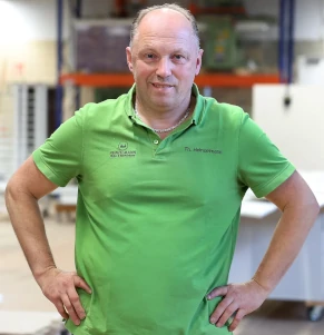 Handwerker in grüner Arbeitskleidung steht in moderner Werkstatt und blickt in die Kamera.