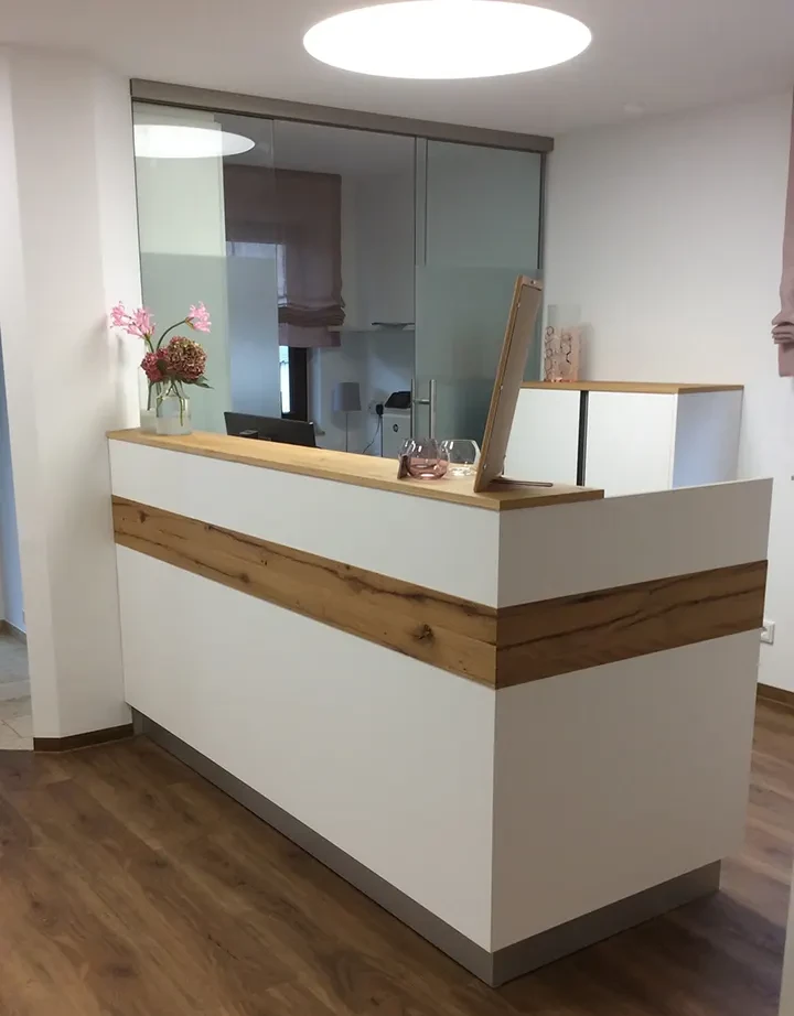 Moderner Empfangstresen aus Holz in heller Praxis mit Glaswand und Dekoration.